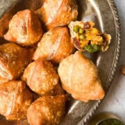 Crunchy veg potato and peas samosas on a platter served with cilantro mint chutney in a small cup.